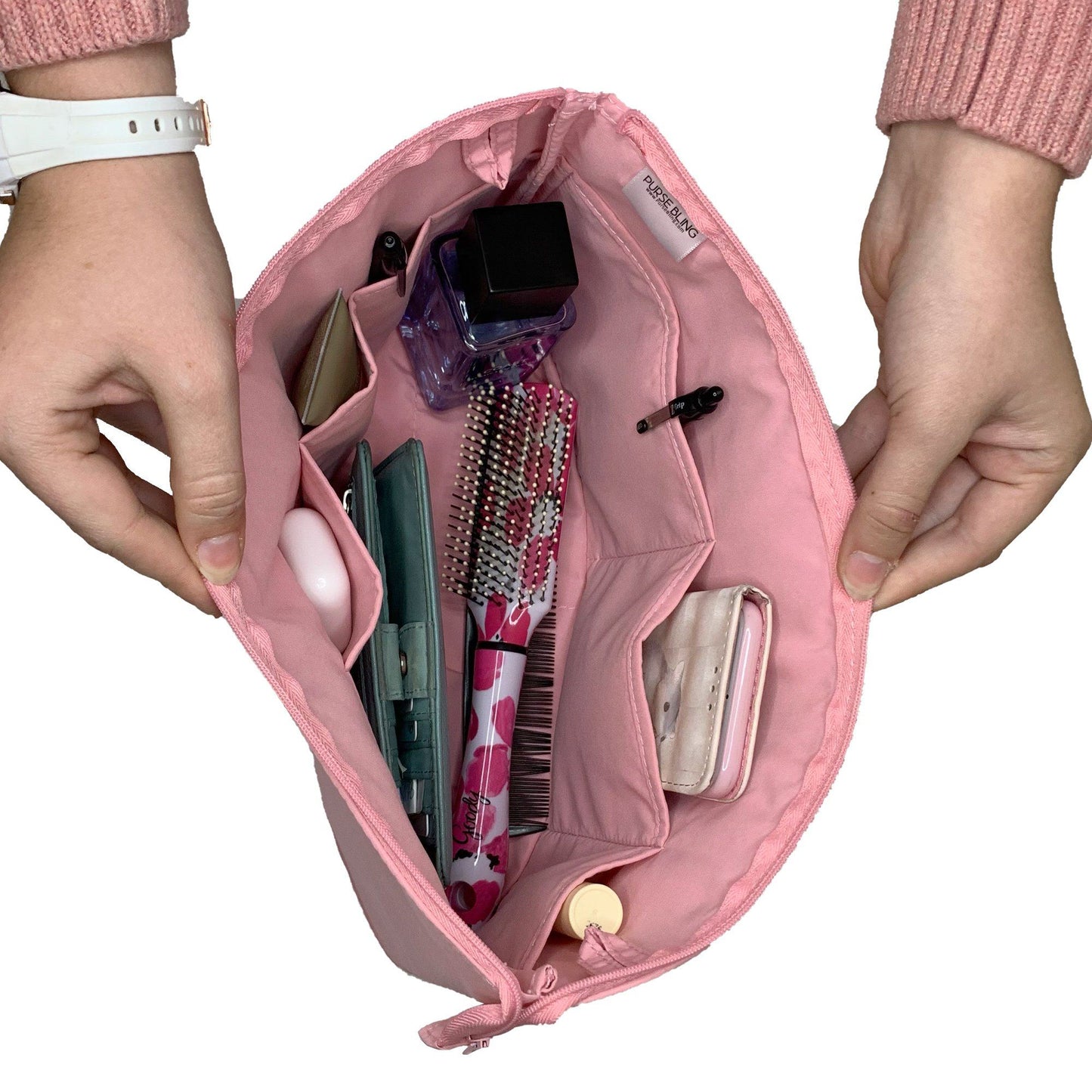 Hands holding the Purse Bling Slim Purse Organizer Insert with Zipper - Extra Jumbo in pink, crafted from polyester. It's filled with personal items like a hairbrush, perfume, wallet, keys, and a pen, all securely tucked inside thanks to its high-quality zipper.